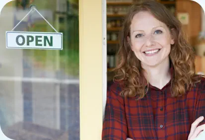 A young woman entrepreneur smiles and leans against the door of her shop which displays an "open" sign